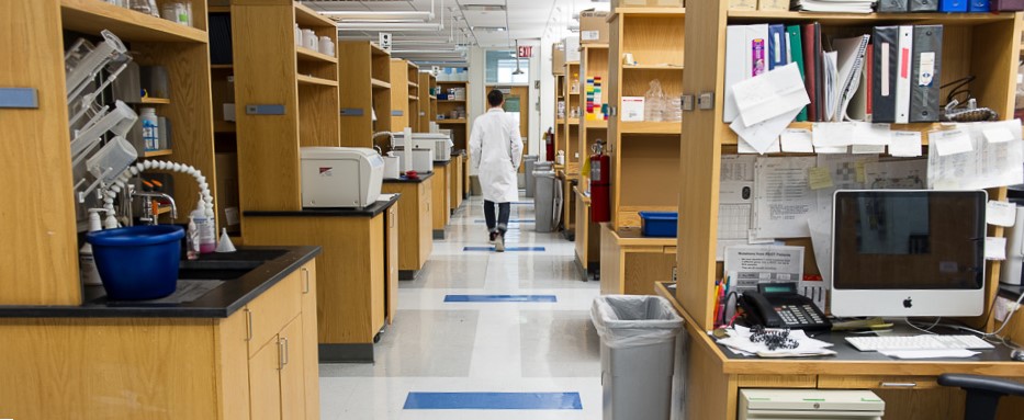Back of man in white coat walking down lab aisle.