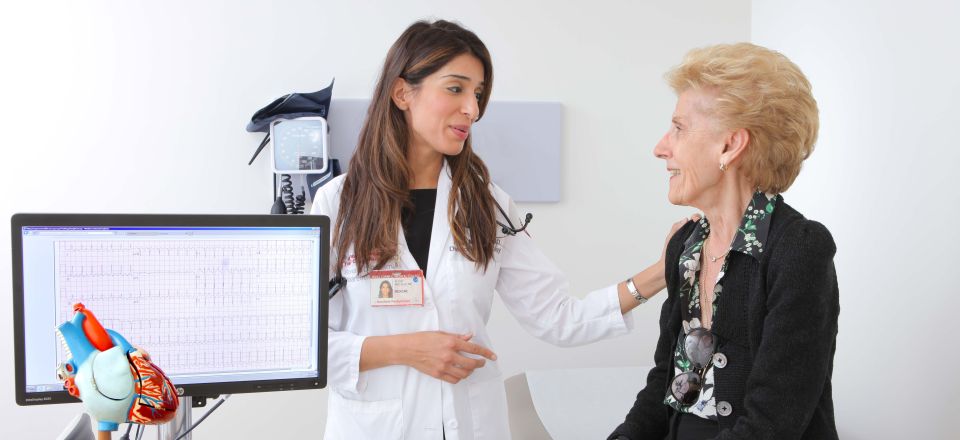 Dr. Alicia Mecklai speaking with female patient featuring ECG and model of heart on table. 